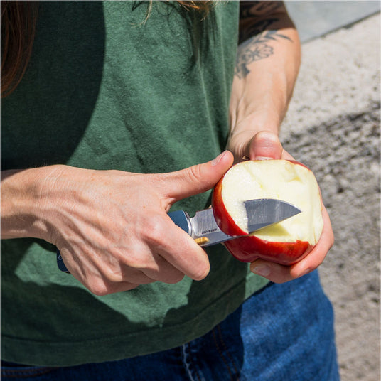 Translation missing: en.Person using a Leatherman Blazer folding knife to peel an apple outdoors, shown in a close-up with the knife blade slicing through the apple skin