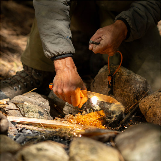 Translation missing: en.Person using a Leatherman Pioneer knife and fire starter to ignite feather sticks in a campfire setup, shown in an outdoor setting with sparks and smoke rising from the tinder