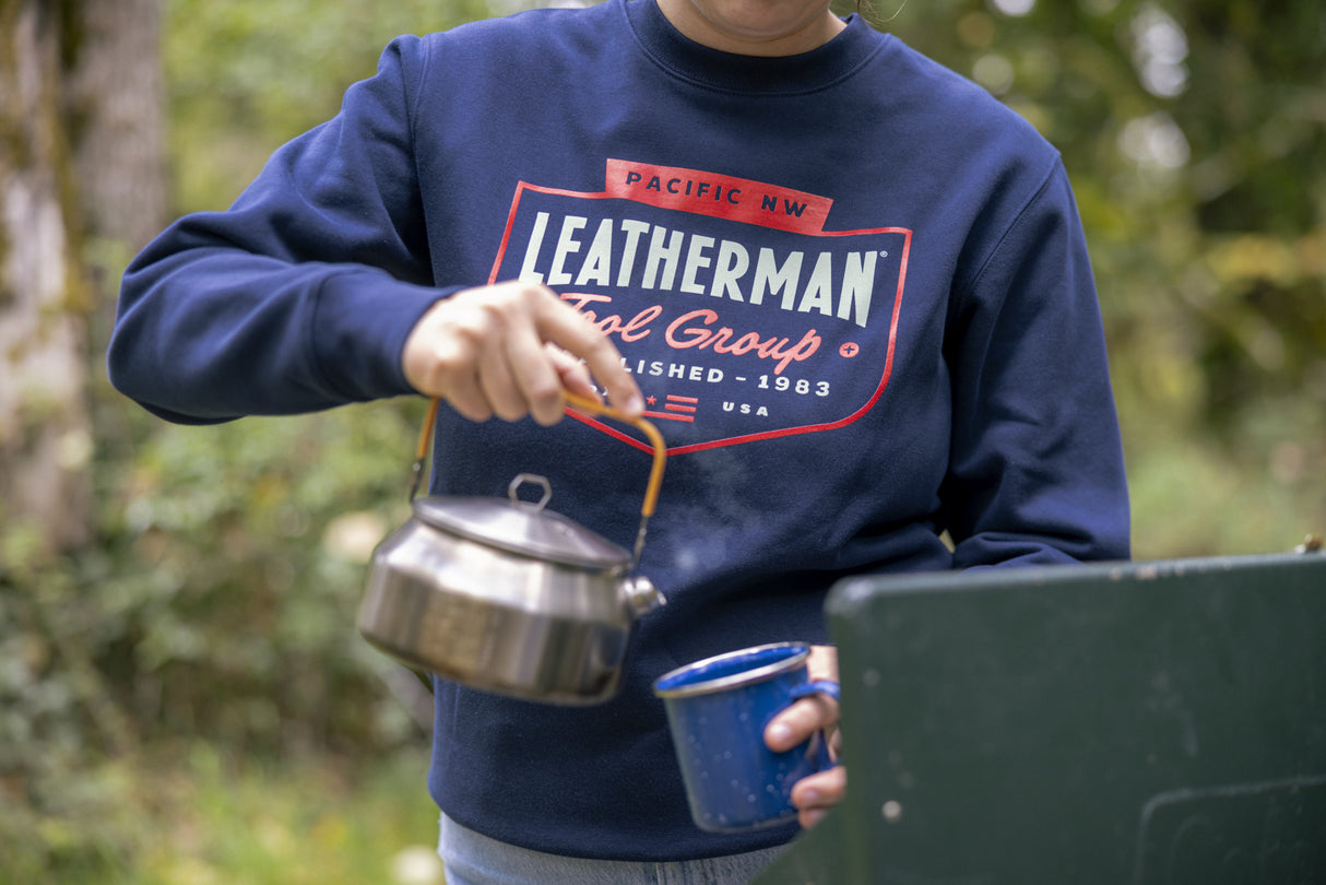 Translation missing: en.Close-up of a person wearing a navy blue Leatherman PNW Crewneck sweatshirt while pouring hot water from a kettle into a blue enamel mug at a campsite