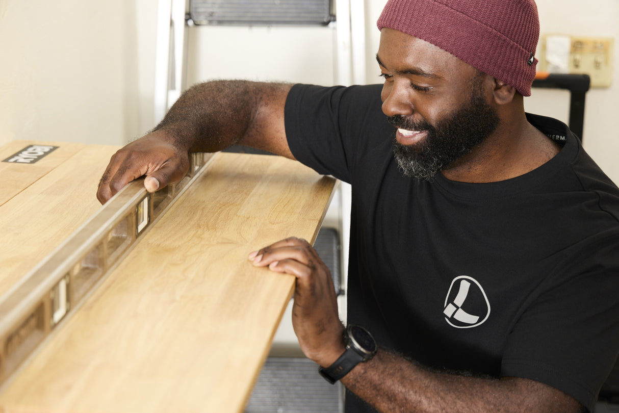 Translation missing: en.A person wearing a black Leatherman Classic Tee using a level tool to level a wooden shelf indoors