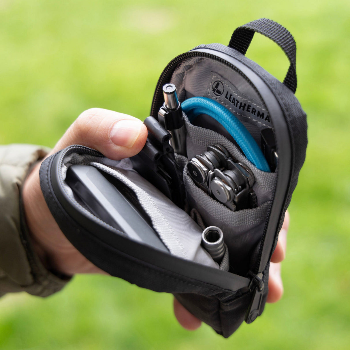 Translation missing: en.A hand holds an open black Leatherman Tool Pouch, revealing a neatly organized interior with a multi-tool, flashlight, pen, charging cable, and other everyday carry essentials. The pouch features a durable design with multiple compartments and a branded interior label