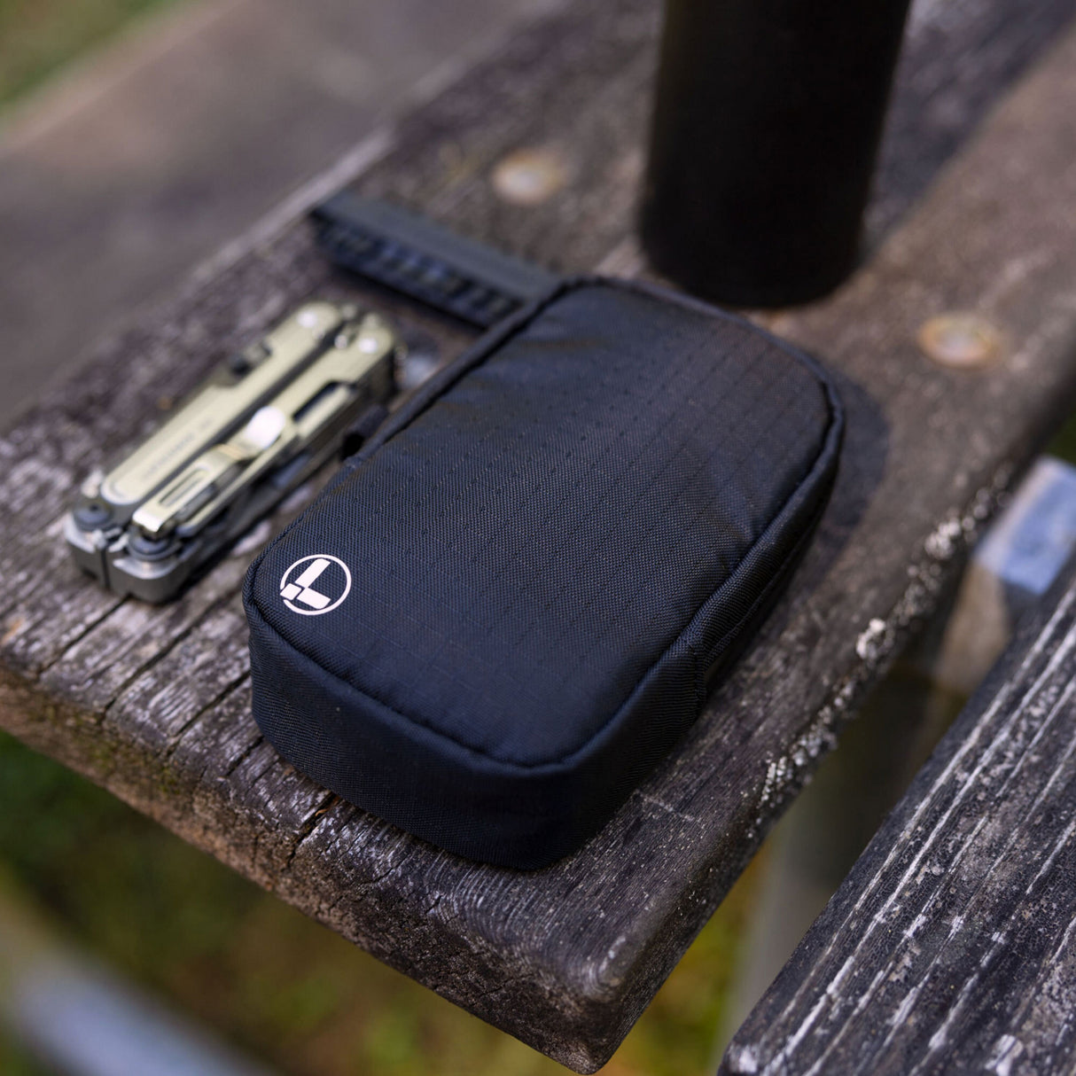 Translation missing: en.A black Leatherman Tool Pouch with a white logo rests on a weathered wooden bench outdoors, positioned beside a Leatherman ARC multi-tool and black water bottle, suggesting a rugged, ready-for-anything everyday carry setup