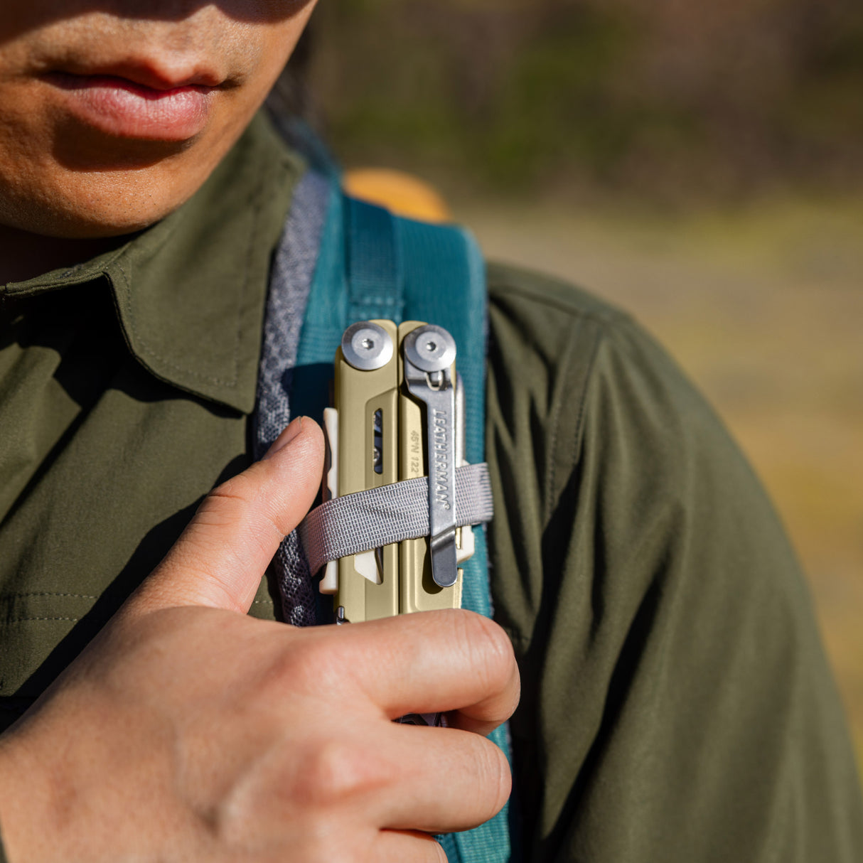Translation missing: en.Person securing a Sandstorm multitool to a backpack shoulder strap with a gray loop outdoors