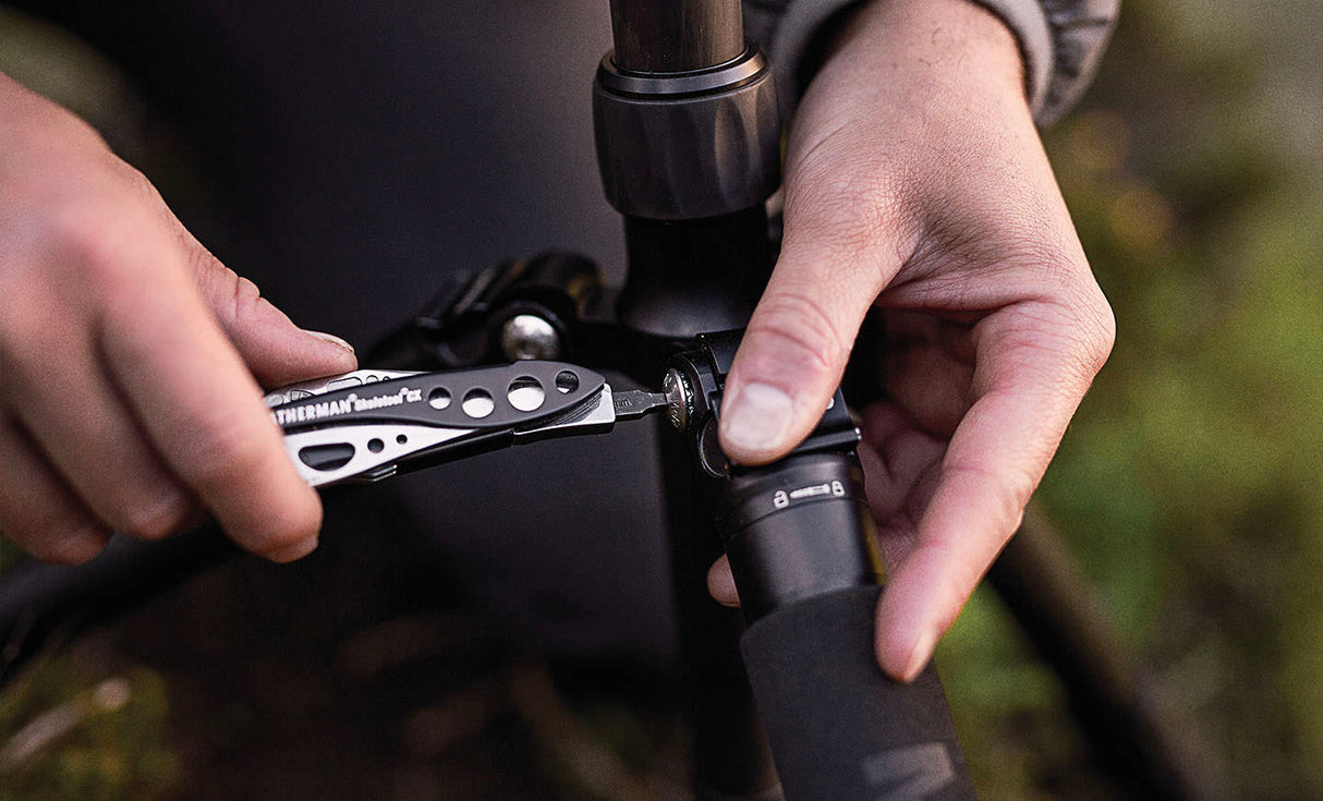 Translation missing: en.Person using a Black & Stainless Leatherman Skeletool® CX multi-tool to adjust a camera tripod in an outdoor setting