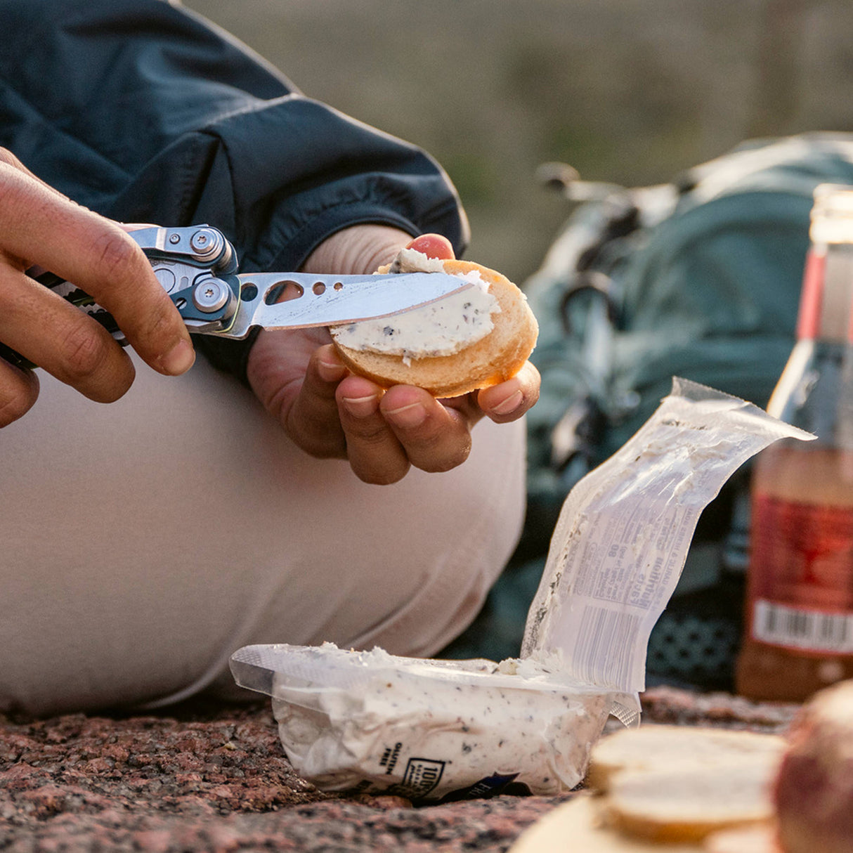 Translation missing: en.Person using a Nightshade Leatherman Skeletool CX multitool knife to spread herbed cheese on a cracker outdoors