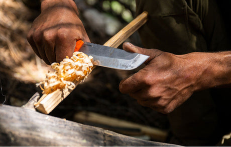 Translation missing: en.Close-up of a person using a Leatherman knife with an orange handle to carve wood shavings from a stick, surrounded by natural outdoor elements, suggesting a bushcraft or survival setting