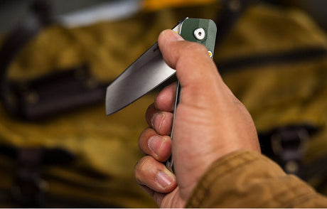 Translation missing: en.Close-up of a person holding a partially open Leatherman folding knife with a green handle, positioned in front of a yellow canvas bag with leather straps