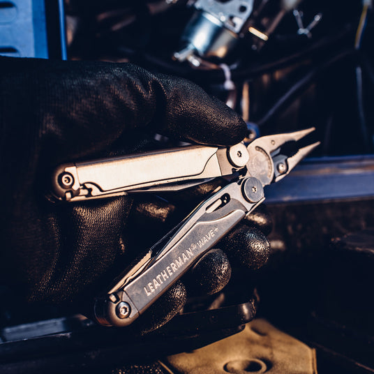 Translation missing: en.A person wearing black gloves holding a Leatherman Wave+, showcasing it&#39;s pliers and tools inside the handle, getting ready to work on a vehicle