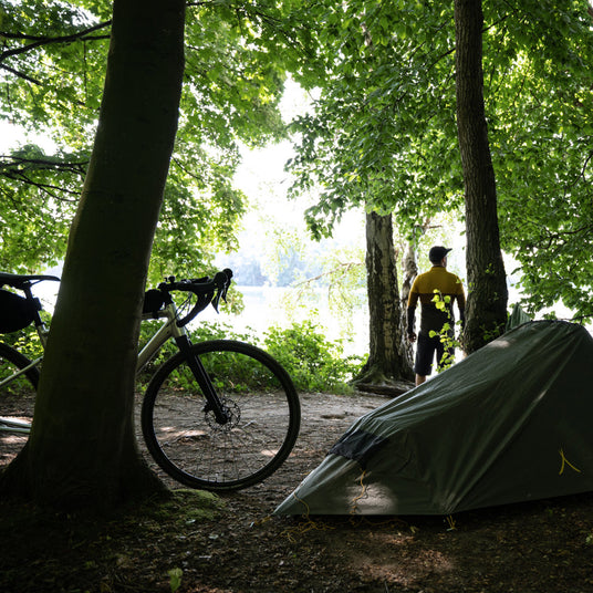 Cyclist standing beside a tent and bicycle in a forest clearing near a lake