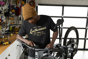 Translation missing: en.A person wearing a black Leatherman Vintage Tee working on a mountain bike in a garage workshop, surrounded by tools and equipment
