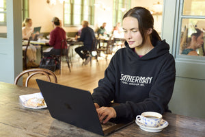 Translation missing: en.A woman sitting indoors working on a computer while wearing a black Leatherman Blade &amp; Grain hoodie with a pastry and coffee on either side of the laptop