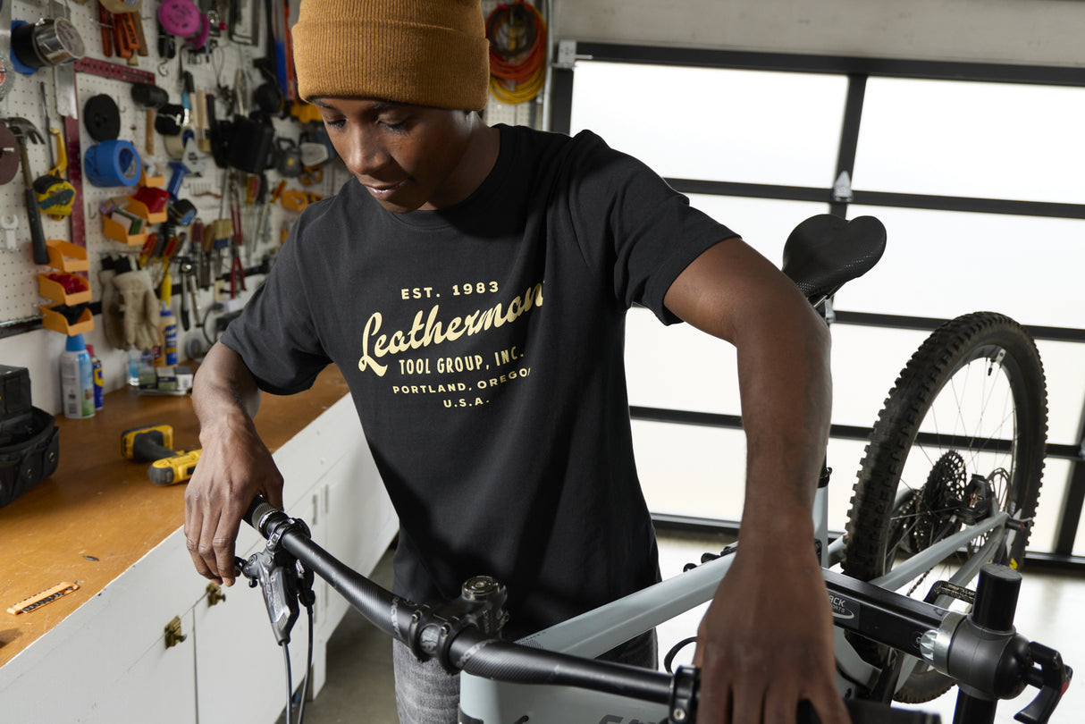 Translation missing: en.A person wearing a black Leatherman Vintage Tee working on a mountain bike in a garage workshop, surrounded by tools and equipment