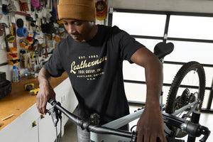 Translation missing: en.A person wearing a black Leatherman Vintage Tee working on a mountain bike in a garage workshop, surrounded by tools and equipment