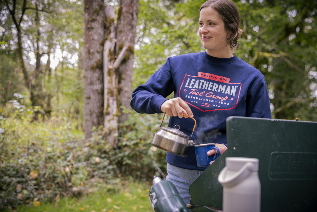 Translation missing: en.A person wearing a navy blue Leatherman PNW Crewneck sweatshirt pouring hot water from a kettle into a mug at a campsite surrounded by trees