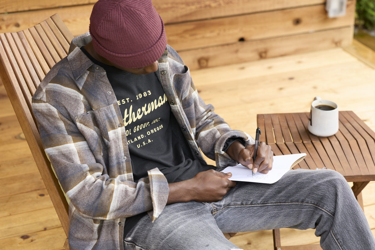 Translation missing: en.A person wearing a black Leatherman Vintage Tee sitting on a wooden chair outdoors, writing in a notebook with a mug of coffee on a nearby table