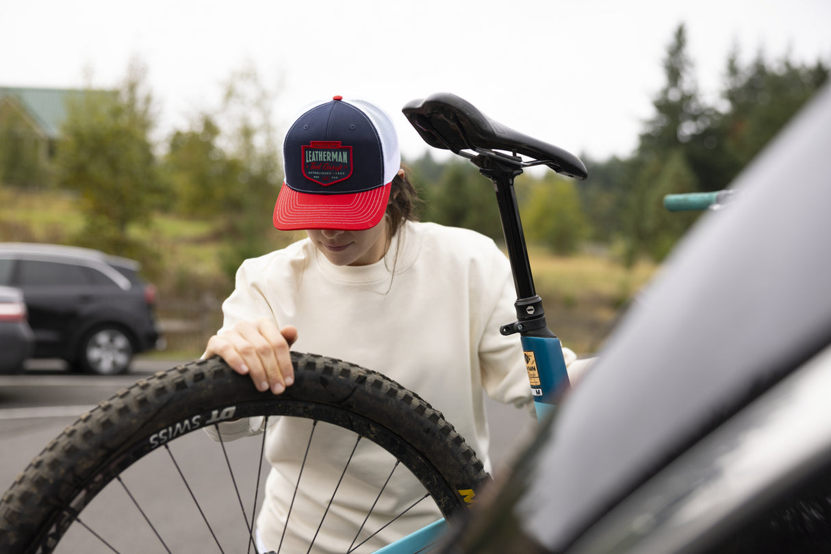 Translation missing: en.A person wearing a navy Leatherman PNW Trucker Hat with a white mesh back and red brim loading a blue mountain bike onto a vehicle