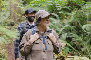 Translation missing: en.A woman wearing a Leatherman Dad Hat in Driftwood color hiking through a forest with a backpack smiling as another hiker follows behind