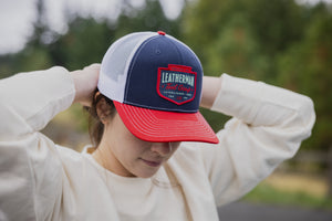 Translation missing: en.A person adjusting a navy Leatherman PNW Trucker Hat with a white mesh back and red brim while outdoors