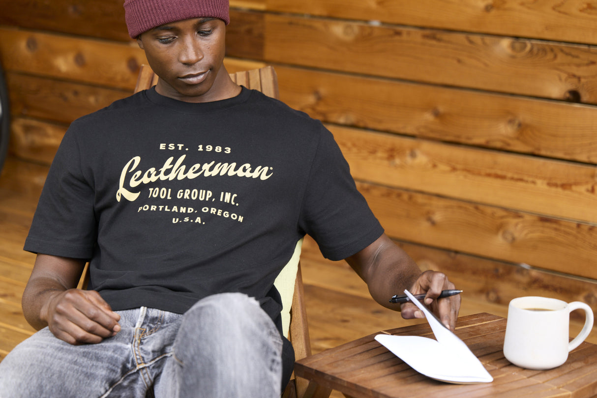 Translation missing: en.A person wearing a black Leatherman Vintage Tee sitting on a wooden deck chair, opening a notebook beside a mug of coffee