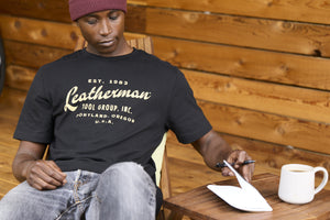 Translation missing: en.A person wearing a black Leatherman Vintage Tee sitting on a wooden deck chair, opening a notebook beside a mug of coffee