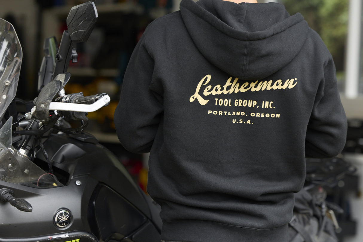 Translation missing: en.A person wearing a black Leatherman Vintage Zip-Up Hoodie standing next to a motorcycle, showing the gold Leatherman Tool Group, Inc. logo printed on the back