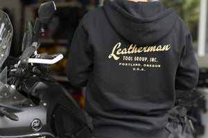 Translation missing: en.A person wearing a black Leatherman Vintage Zip-Up Hoodie standing next to a motorcycle, showing the gold Leatherman Tool Group, Inc. logo printed on the back