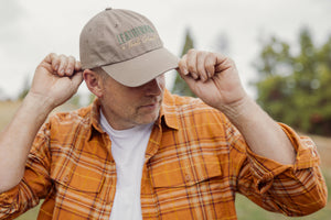 Translation missing: en.A person wearing a Leatherman Dad Hat in Driftwood color adjusting the brim while outdoors, dressed in an orange plaid shirt