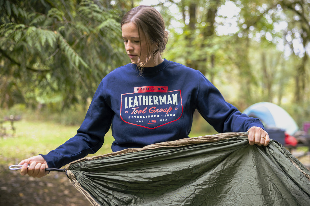 Translation missing: en.A person wearing a navy blue Leatherman PNW Crewneck sweatshirt setting up a hammock at a forest campsite with a tent in the background