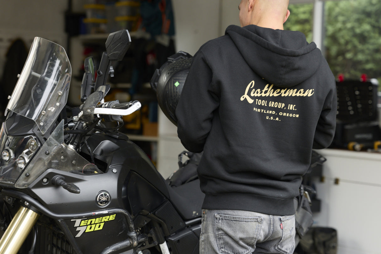 Translation missing: en.A person wearing a black Leatherman Vintage Zip-Up Hoodie standing next to a motorcycle, showing the gold Leatherman Tool Group, Inc. logo printed on the back