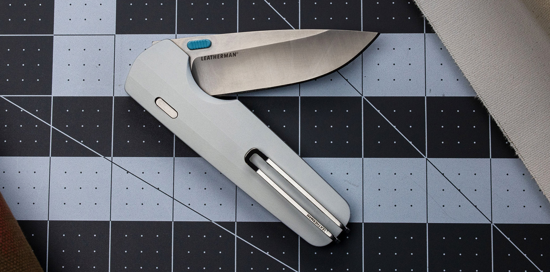 Leatherman Blazer Stainless folding knife shown partially open on a gridded cutting mat, next to a cloth canvas, highlighting its utility in crafting or workshop environments.