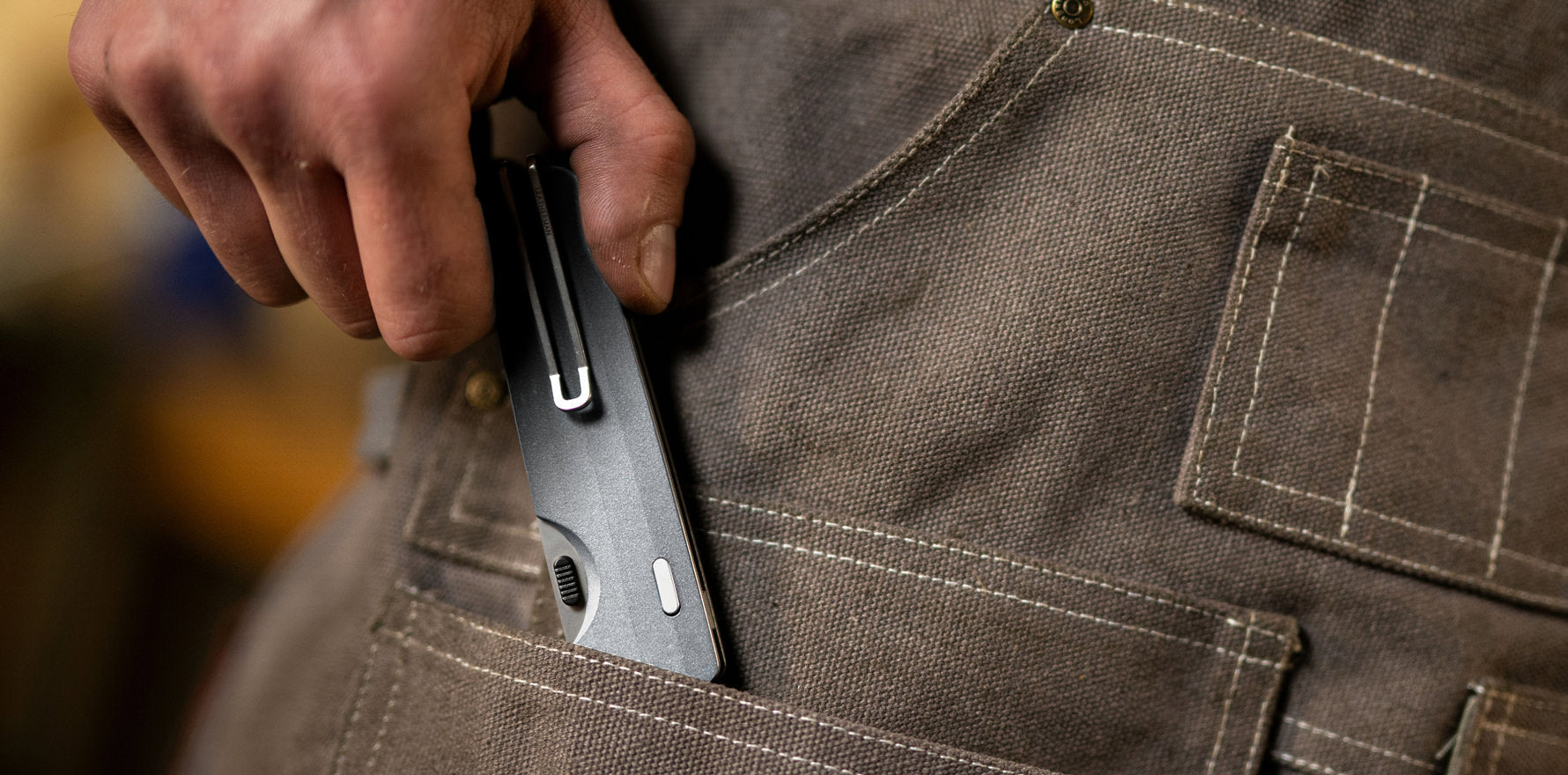 Person sliding a closed Leatherman Blazer folding knife into the pocket of a rugged canvas work apron, emphasizing practical everyday carry for workshop or field use.