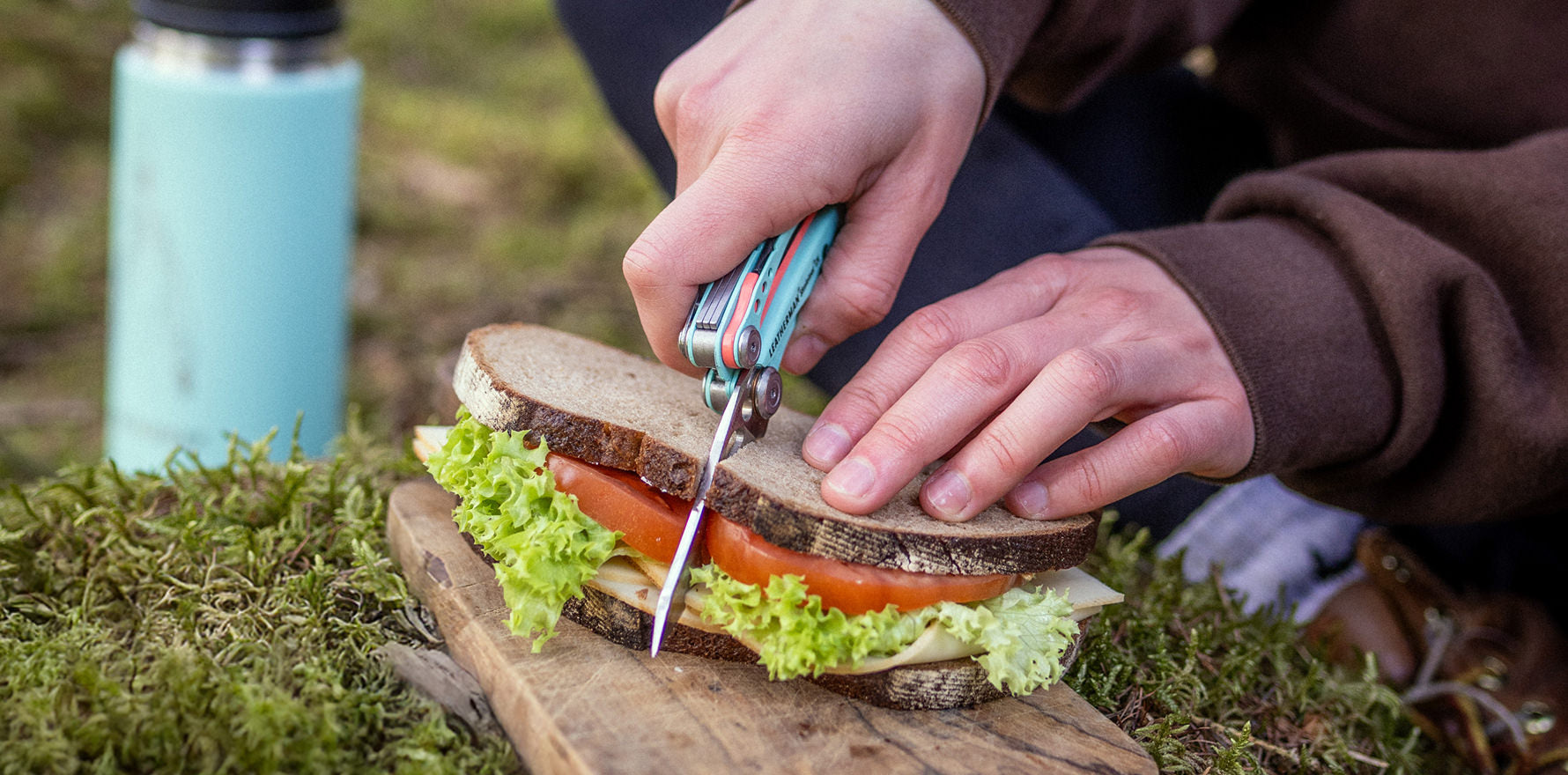 A hand using a Paradise Skeletool CX to slice a sandwich next to a Yeti bottle outdoors.