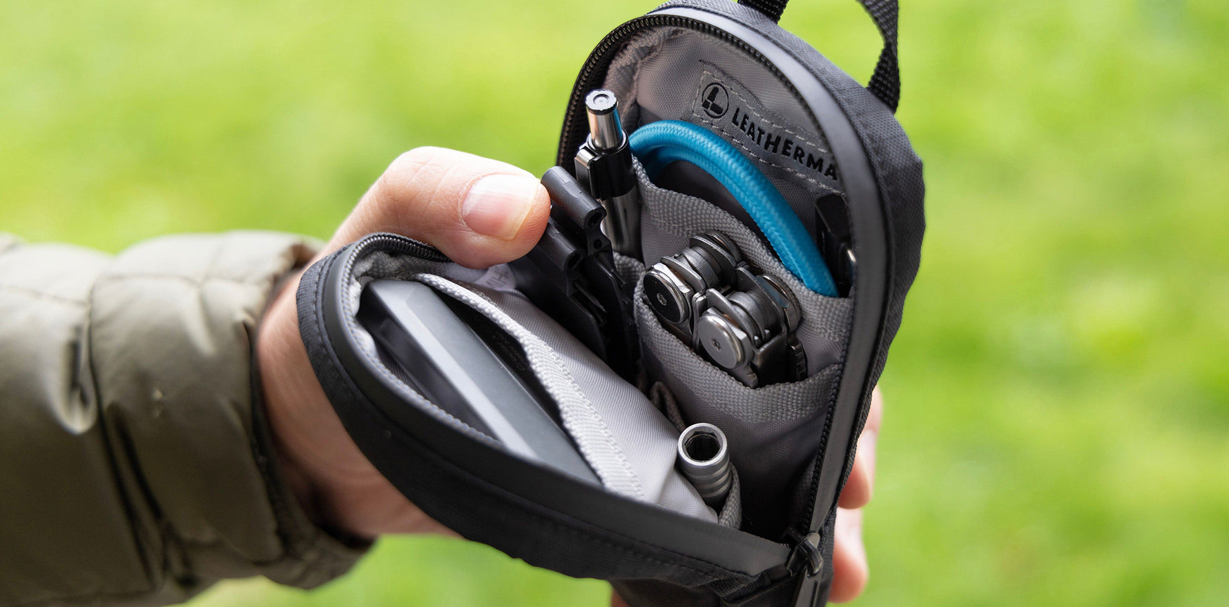 A person holds open a Leatherman Tool Pouch in black color, revealing a neatly organized interior containing a multitool, bit kit, pen, cables, and other everyday essentials, with a blurred green outdoor background.