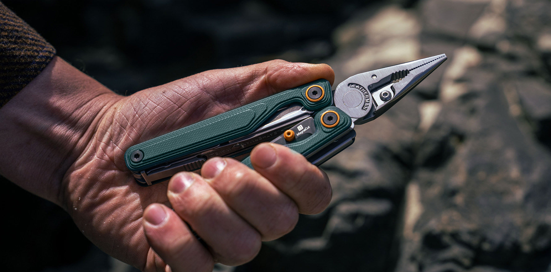 Hand holding the Leatherman Wave® Alpha Cascadia multi-tool outdoors, pliers open against a blurry rocky background.
