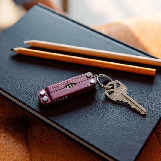 Closed Leatherman Cherry Micra on keyring with key, above black notebook, next to 2 yellow pencils. 