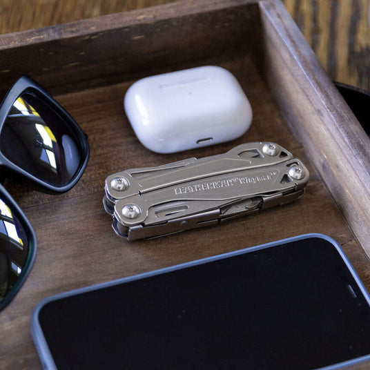 Closed Leatherman Wingman in wooden box next to airpods, sunglasses and cellular phone. 