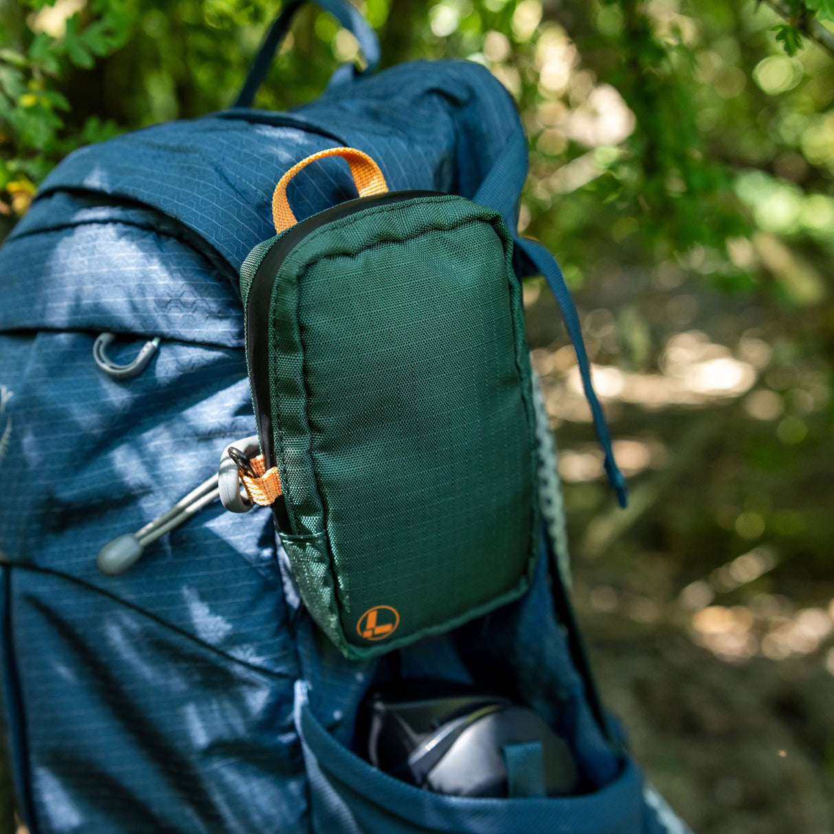 Translation missing: en.A Leatherman Tool Pouch in Hunter color is clipped to the shoulder strap of a blue hiking backpack, resting in an outdoor forest setting with sunlight filtering through the trees