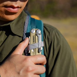 Translation missing: en.Person securing a Sandstorm multitool to a backpack shoulder strap with a gray loop outdoors
