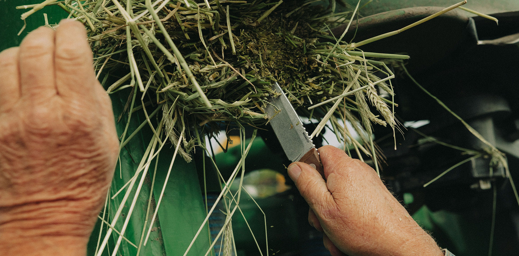 Person using a Leatherman Trac Earth Brown knife to cut through a bundle of hay or straw attached to green agricultural equipment in an outdoor farm setting.