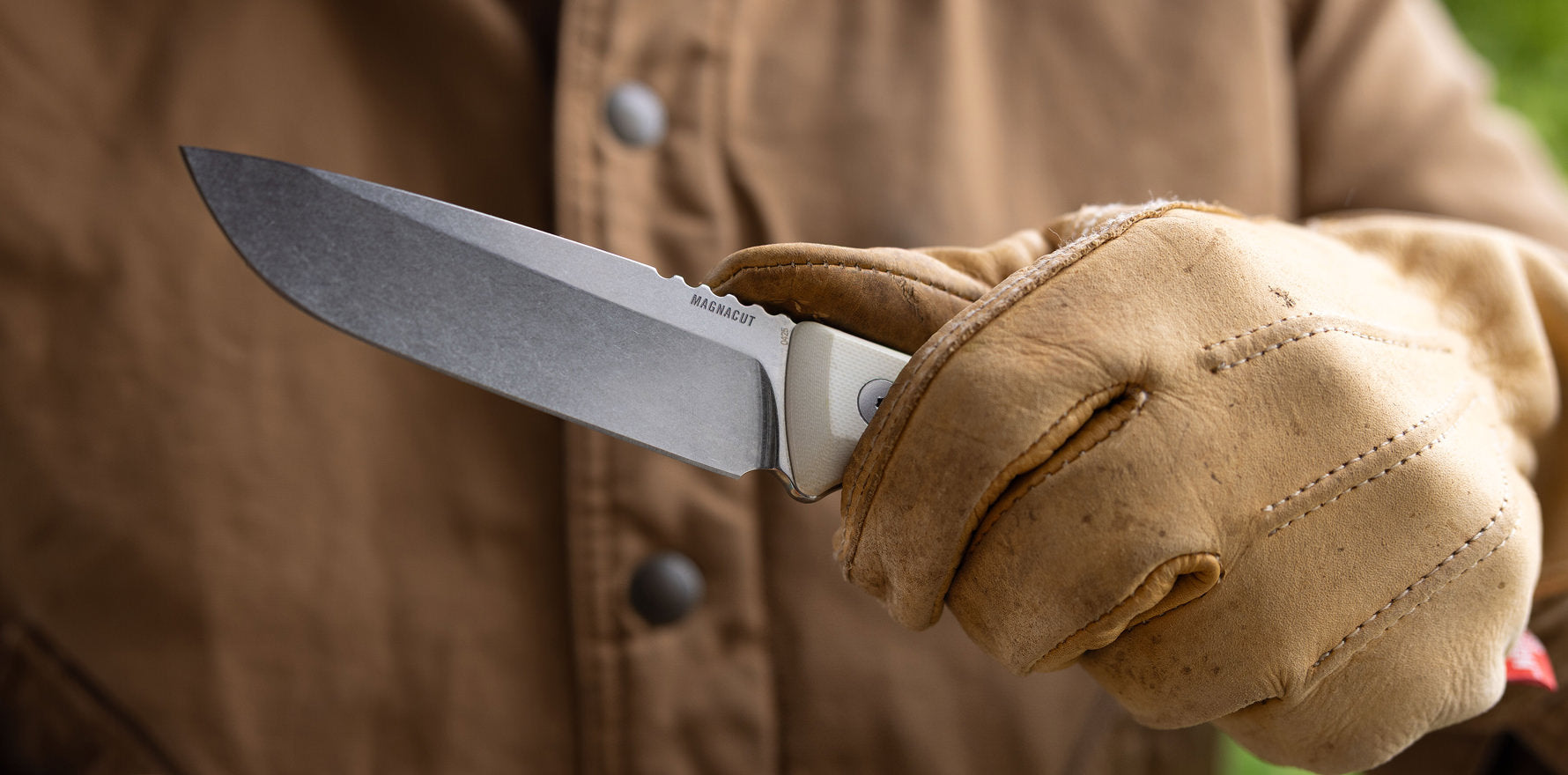 Close-up of a gloved hand holding a Leatherman Trac Alpine knife outdoors, with a focus on the stonewashed blade and white handle against a brown work jacket background.