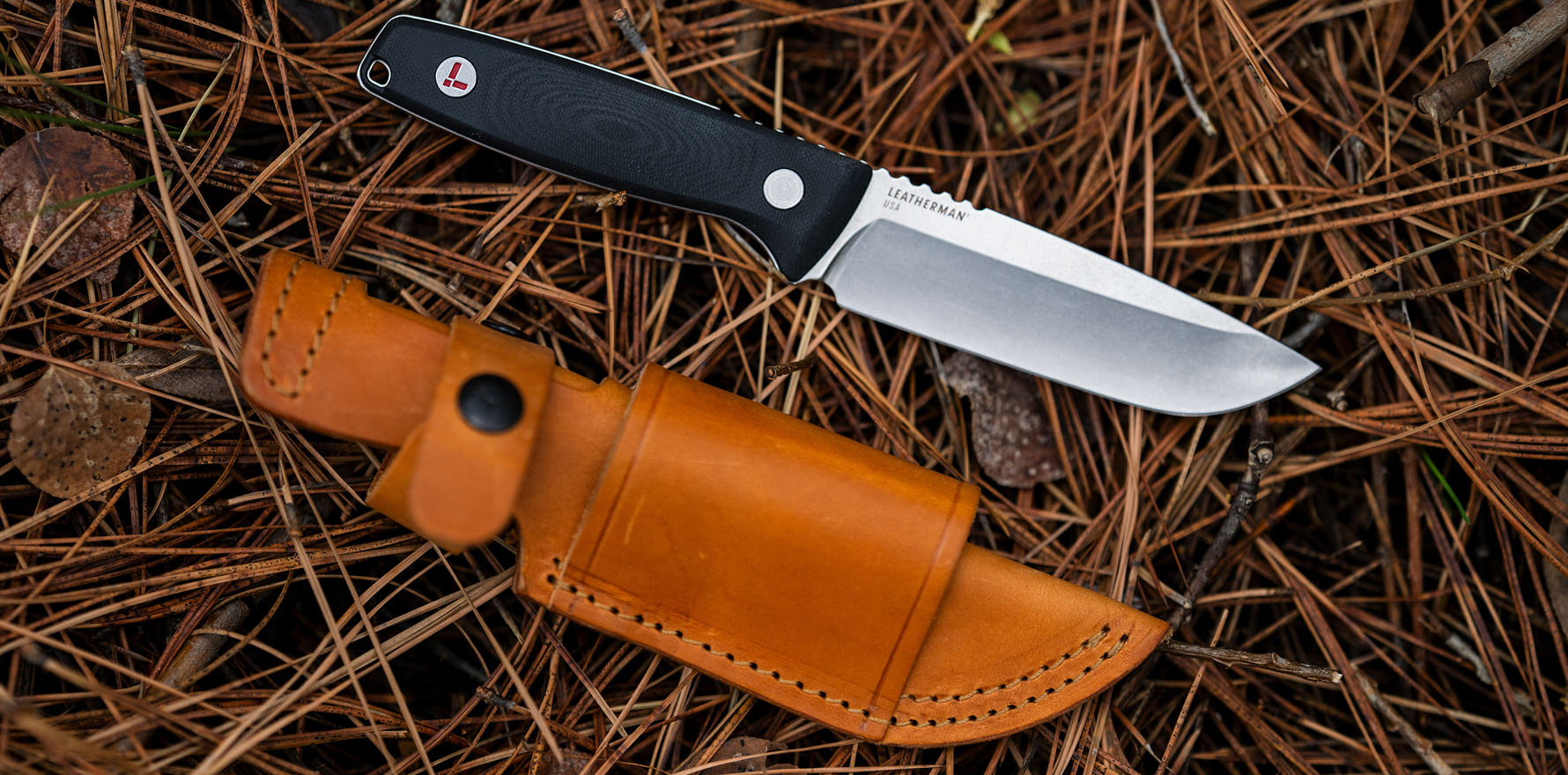 Leatherman Trac Jet Black knife resting on a forest floor covered in pine needles, placed beside a tan leather sheath with black button closure.