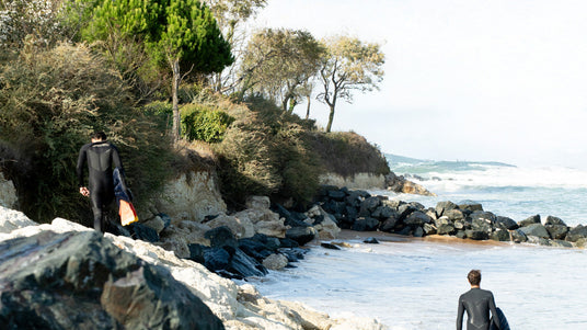 Translation missing: en.Two people in wetsuits walking along a rocky beach with trees and ocean in the background